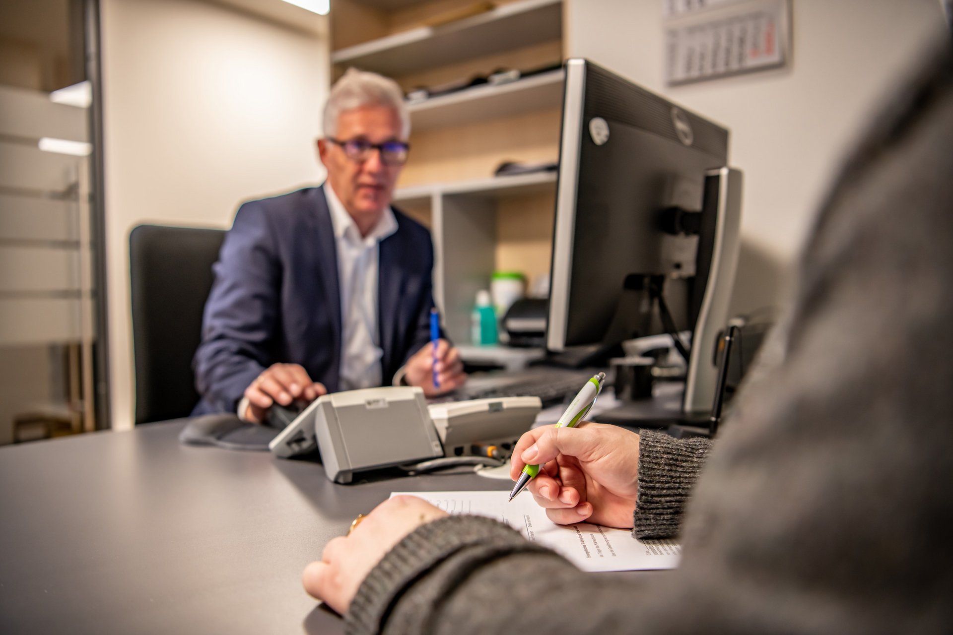 ein Patient beim Ausfüllen eines Formulars und ein Sachbearbeiter in einem Büro